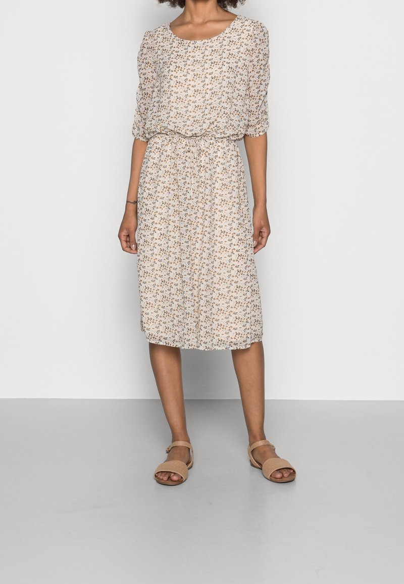 Vrouw in een knie-lengte, witte bloemenjurk met driekwartmouwen en beige open teen sandalen, staande tegen een effen achtergrond.