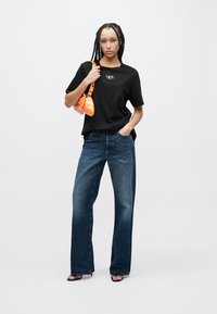 Μαύρο μπλουζάκι με λογότυπο, μπλε φαρδιά τζιν παντελόνια και πορτοκαλί τσάντα. Το μοντέλο φοράει μαύρα σανδάλια και έχει πλεξούδες στα μαλλιά.