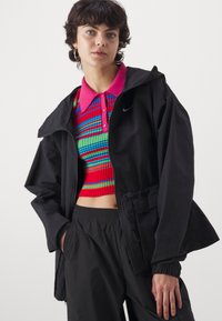 Sort letvægtsjakke med hætte, parret med en lys, flerfarvet stribet cropped top og sorte bukser med elastisk talje.