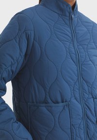 Veste bleue matelassée avec un col haut, présentant des motifs en vagues, un zip sur le devant et deux poches avant. Le matériau semble léger et lisse.