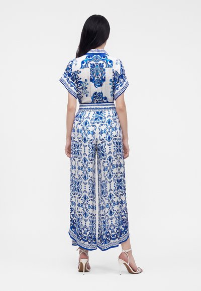 Femme aux longs cheveux foncés portant un haut à manches courtes blanc et bleu à motifs et un pantalon à jambes larges avec des sandales blanches à talons hauts, debout de dos.
