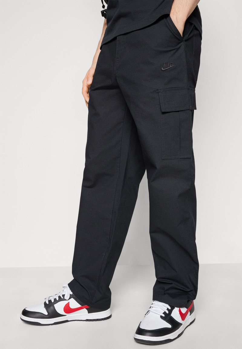 Pantalons cargo noirs au fini texturé, avec des poches latérales et un logo Nike discret sur la cuisse gauche. Accompagnés de baskets noires et blanches.