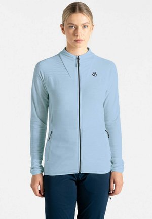 Junge Frau, die nach vorne blickt, trägt eine hellblaue Reißverschlussjacke und dunkle Hose, steht vor einem einfachen weißen Hintergrund.