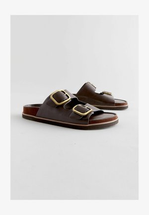 Bruine leren sandalen met twee brede banden, elk met een grote gouden gesp, en een platte zool met een lichte bruine rand.