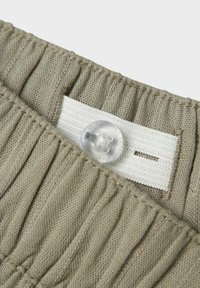 Fascia elastica in tessuto khaki con pieghe verticali, dotata di un'etichetta bianca e un dettaglio con bottone trasparente. Materiale leggero e testurizzato.