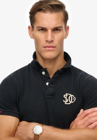 Schwarzes Poloshirt mit kurzem Kragen, zwei Knöpfen und einem bestickten Logo in Creme. Das Modell trägt eine braune Uhr und hat die Arme verschränkt.