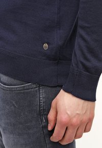 Granatowy sweter wykonany z miękkiego materiału, z prążkowanymi mankietami i dołem; posiada małą metalową metkę z logo na dolnej stronie.
