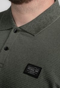 Olijfgroene polo met een gestructureerde stof. Bevat drie zwarte knopen en een zwart vierkant logo op de borst.