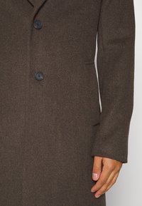 Cappotto in misto lana marrone con due bottoni neri, colletto a punta e tasche laterali. La texture liscia e la forma sartoriale esaltano il suo aspetto strutturato.