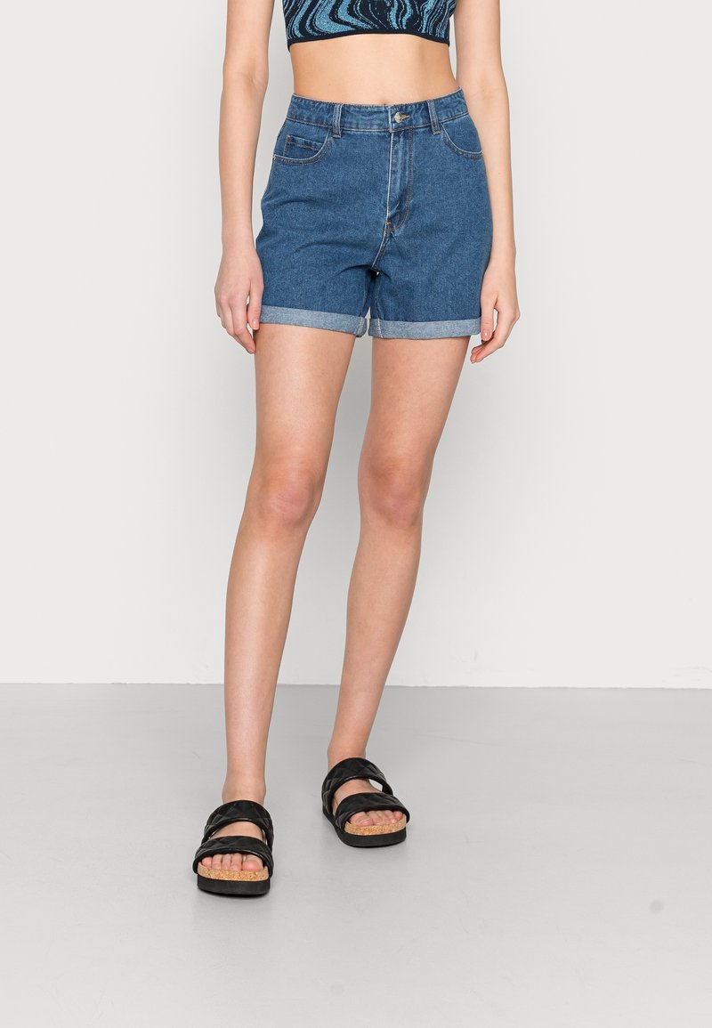 Džínové šortky v středně modré barvě s ohrnutými lemy, obsahující pět kapes a zapínání na knoflík, ve spojení s černými sandály na pásky.