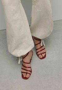 Red strappy sandals with flat soles, featuring narrow straps and a matte finish. Worn with loose, light-colored pants. Smooth textured floor.