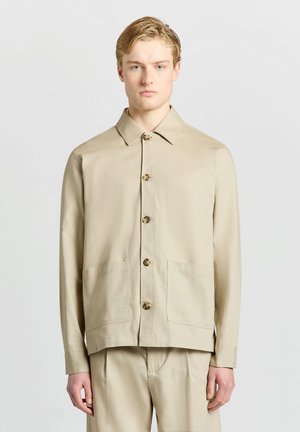 Junger Mann mit hellem Haar, der eine beige Jacke mit Knöpfen und Kragen sowie passende beige Hose vor einem schlichten hellen Hintergrund trägt.