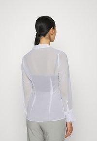 Camisa blanca, transparente, de mangas largas con cuello clásico y puños abotonados. Presenta un corte entallado y una textura suave.