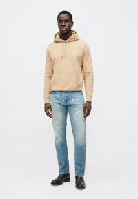 Μπεζ φούτερ με κουκούλα και μπροστινή τσέπη, σε συνδυασμό με ανοιχτό μπλε τζιν με φθορές. Το μοντέλο φοράει μαύρα μποτάκια αστραγάλου, στέκεται μπροστά σε απλό φόντο.