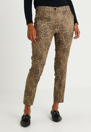 Nainen, joka pukeutunut kapeisiin leopardikuvioisiin housuihin, mustaan neulepaitaan ja mustiin hapsullisiin loafereihin, seisoo kädet rentoina sivuilla.