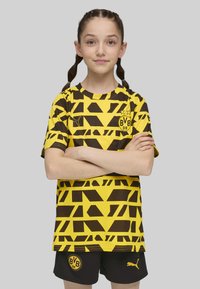 Camiseta de fútbol amarilla y marrón con patrones geométricos, de mangas cortas y cuello redondo, combinada con pantalones cortos negros que tienen un logo.