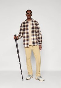 Rudos, kreminės ir tamsiai mėlynos spalvų languota flanelinė palaidinė, vilkima ant smėlio spalvos marškinėlių, derinama su smėlio spalvos kelnėmis ir pilkais sportbačiais. Laiko juodą žygių lazdą.