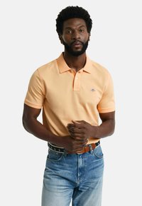 Kortermet polo-skjorte med krage i lysoransje stoff, med en liten blå logo. Brukt med blå denimjeans og et brunt belte.