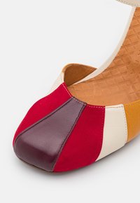 Gros plan d'une chaussure plate multicolore avec des panneaux en cuir rouge, bordeaux, crème et moutarde et une semelle intérieure texturée beige.