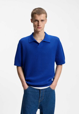 Junger Mann mit kurzen Haaren, der einen strukturierten königsblauen Kurzarm-Polo-Pullover und blaue Jeans trägt, steht mit den Händen in den Taschen.