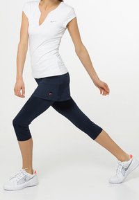 Camiseta blanca de cuello en V combinada con una falda azul marino, leggings capri ajustados azul marino y zapatillas blancas con detalles en naranja, en un modelo en movimiento.