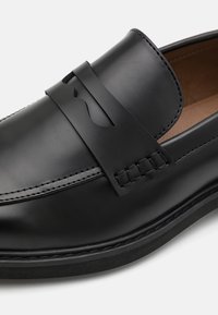 Schwarzer Lederloafer mit einem eleganten Design, das einen dekorativen Riemen und eine Nahtdetail aufweist, sowie einer strukturierten schwarzen Gummisohle.