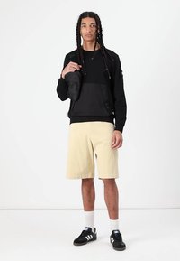 Svart langermet skjorte med lomme, lysfargede shorts, svarte joggesko og hvite sokker; stående posisjon mot en enkel bakgrunn.