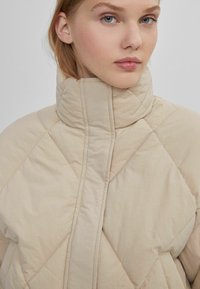 Beige quiltet jakke med høj krave, der har diamantmønstre og en glat tekstur. Lynlås i front og minimalistisk design.