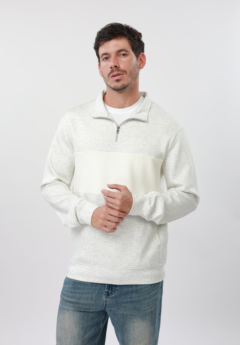 Homme aux cheveux courts et foncés, portant un sweat-shirt gris clair à fermeture éclair quart et un jean bleu, debout devant un fond blanc uni.