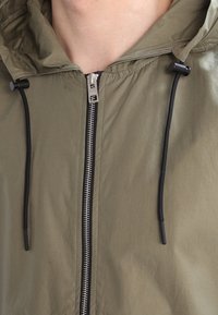 Gros plan sur une personne portant une veste à capuchon vert olive avec une fermeture éclair argentée et des cordons de serrage noirs.