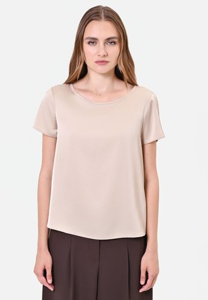 Donna con capelli lisci castano chiaro indossa una maglietta beige a maniche corte e pantaloni plissettati marrone scuro, in piedi davanti a uno sfondo bianco semplice.