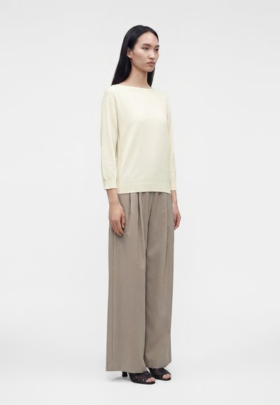 Femme aux longs cheveux foncés debout, portant un haut crème à manches longues, un pantalon beige large et des talons ouverts noirs, sur fond blanc.