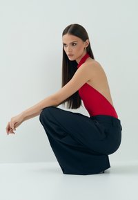 Czerwony body na halter z odkrytymi plecami, zestawione z szerokimi granatowymi spodniami; gładki materiał i minimalistyczny design. Modelka przyjmuje postawę kucającą.