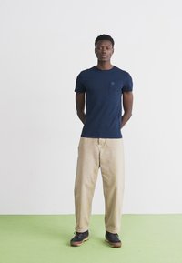 Cămașă de tip t-shirt bleu marin cu mânecă scurtă, dotată cu un buzunar pe piept, asociată cu pantaloni largi de culoare khaki și pantofi închisi la culoare, stând pe un podea verde.
