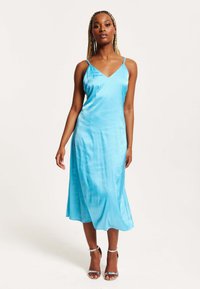 A satin, turquoise slip dress with a V-neck and thin straps, featuring a flowy silhouette that falls mid-calf. Silver heeled sandals.