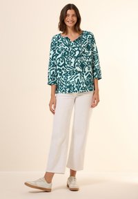 Blusa stampata turchese e bianca con scollo rotondo, maniche a 3/4, abbinata a pantaloni bianchi a gamba larga e scarpe da ginnastica chiare.
