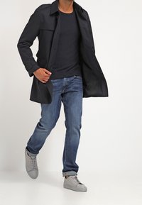 Černý trenčkot přes tmavý svetr, doplněný modrými džínami a světle šedými teniskami. Má límec a zapínání na knoflíky na přední straně.