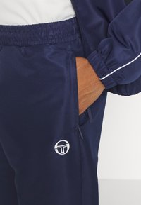 Navy athletic tracksuit bottoms featuring a textured fabric, elastic waistband, side pockets, and a white logo detail on the left thigh.