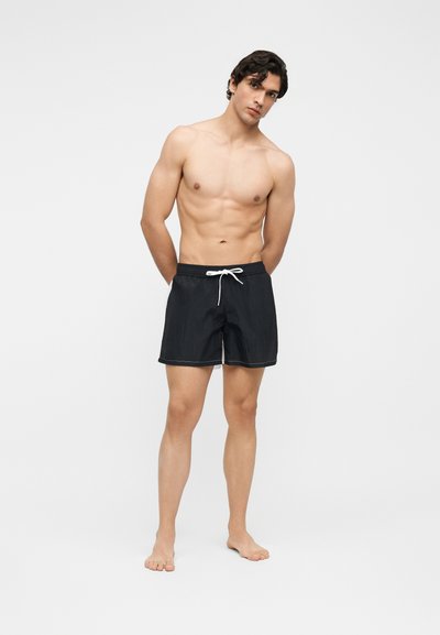 Νέος άνδρας στέκεται ξυπόλητος, χωρίς μπλουζάκι, φορώντας μαύρο μαγιό με λευκό κορδόνι σε απλό ανοιχτό φόντο.