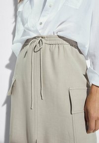 Hvid skjorte med knapper, der er stukket ind i en beige nederdel med snørebånd og store påsyede lommer, vist fra talje til midt på låret på en person.