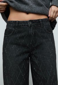 Czarne dżinsy z denimu z ozdobami z diamentowego wzoru w kamieniach szlachetnych. Posiadają zapięcie na guzik i tradycyjne kieszenie. Teksturowany materiał.
