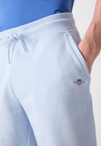 Homme portant un pantalon de survêtement bleu clair avec taille à cordon et main dans la poche, montrant le logo GANT brodé sur la cuisse.