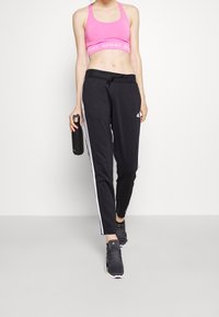 Frau trägt pinken Adidas-Sport-BH und schwarze Adidas-Trainingshose, hält eine schwarze Wasserflasche und steht vor einem weißen Hintergrund.
