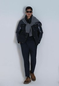 Doudoune marine avec finition brillante, pull gris noué sur les épaules, pantalon sombre et chaussures brunes avec finition texturée. Lunettes de soleil noires.