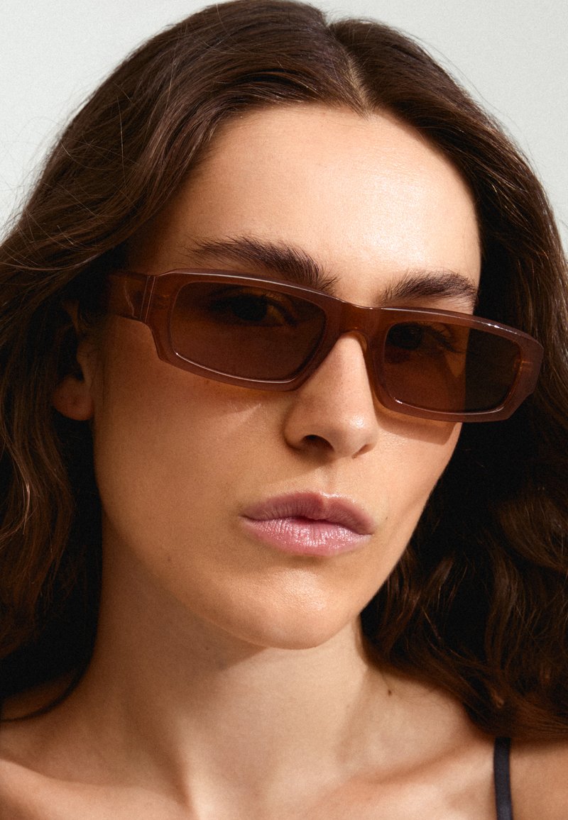 Gafas de sol rectangulares marrones con lentes ahumados, gruesos marcos transparentes y diseño angular. Textura suave con detalles de hardware mínimos.
