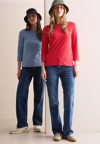 Zwei Models stehen vor einem hellen Hintergrund. Eine trägt ein blau-weiß gestreiftes Oberteil, die andere ein rotes Oberteil mit weißen Streifen, beide in Denim-Jeans.