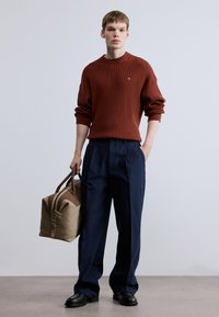 Πλεκτή μπλούζα σε σκουριά, μπλε-ναυτικό παντελόνι με φαρδιά μπατζάκια, μαύρα παπούτσια, κρατώντας μια μπεζ τσάντα duffel με καφέ λεπτομέρειες. Απλός, υφασμάτινος και κομψός εμφανισιακός συνδυασμός.