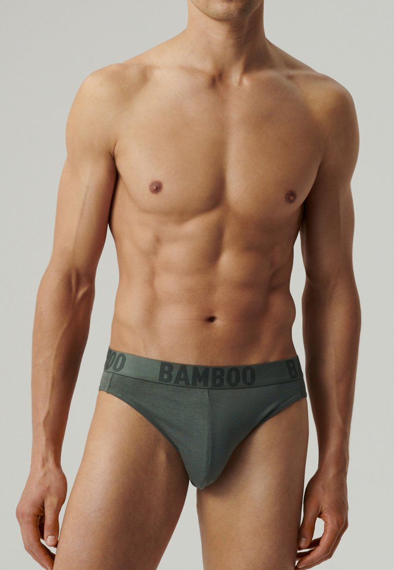 Sprawna męska tors z zielonymi majtkami z logo "BAMBOO" na pasku przeciwko jednolitemu tłu.