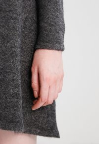 Abito di lana grigia con una trama morbida, dotato di maniche lunghe e un orlo leggermente svasato, che si estende appena sopra la mano.