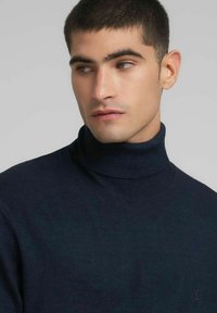 Maglione dolcevita blu navy realizzato in tessuto morbido, presenta un design aderente e un collo a coste. Dettaglio di un logo discreto sul petto.